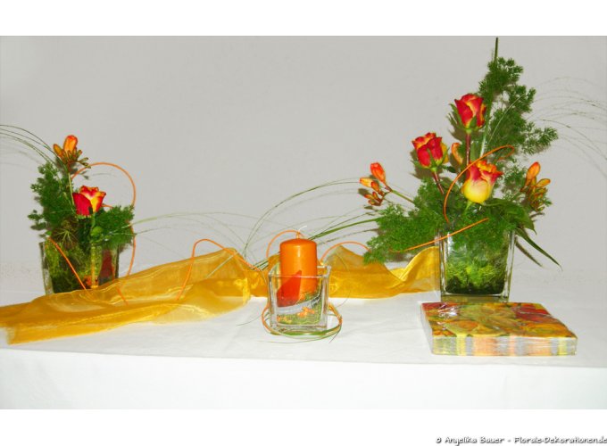 Tischschmuck mit Rosen, Freesien und einer Kerze in drei verschiedenen Gefäßen und farblich passenden Servietten.